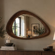 An organic-shaped wooden wall mirror with a dark brown carved frame, mounted on a beige wall above a wooden console table with books and decor.