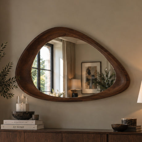 An organic-shaped wooden wall mirror with a dark brown carved frame, mounted on a beige wall above a wooden console table with books and decor.
