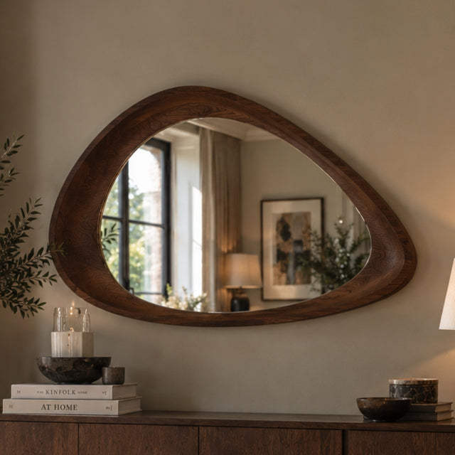 An organic-shaped wooden wall mirror with a dark brown carved frame, mounted on a beige wall above a wooden console table with books and decor.