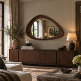 A large organic-shaped wooden wall mirror with a dark wood frame hangs above a mid-century modern wooden credenza in a beautifully styled living room with warm lighting, a table lamp, and decorative plants.