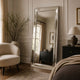 A tall floor mirror with a silver frame stands in a styled bedroom setting, reflecting a window and dresser in the room behind it. The scene includes a cream armchair, a dark wood nightstand with a lamp, and neutral-toned bedding.