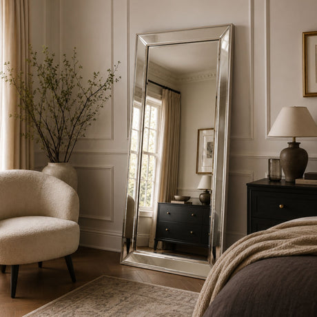 A tall floor mirror with a silver frame stands in a styled bedroom setting, reflecting a window and dresser in the room behind it. The scene includes a cream armchair, a dark wood nightstand with a lamp, and neutral-toned bedding.