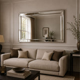 A large rectangular silver-framed mirror hangs on a beige paneled wall above a light beige sofa with dark brown accent pillows in a styled living room.