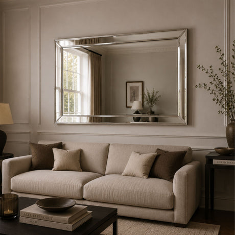 A large rectangular silver-framed mirror hangs on a beige paneled wall above a light beige sofa with dark brown accent pillows in a styled living room.