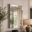 A rectangular wall mirror with a slim silver frame hangs on a beige paneled wall. The mirror reflects a window with cream curtains and greenery, while the surrounding room includes a potted plant on the left and a table lamp on the right.