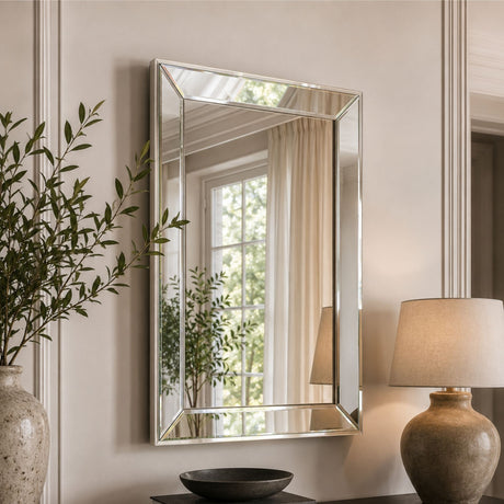 A rectangular wall mirror with a slim silver frame hangs on a beige paneled wall. The mirror reflects a window with cream curtains and greenery, while the surrounding room includes a potted plant on the left and a table lamp on the right.