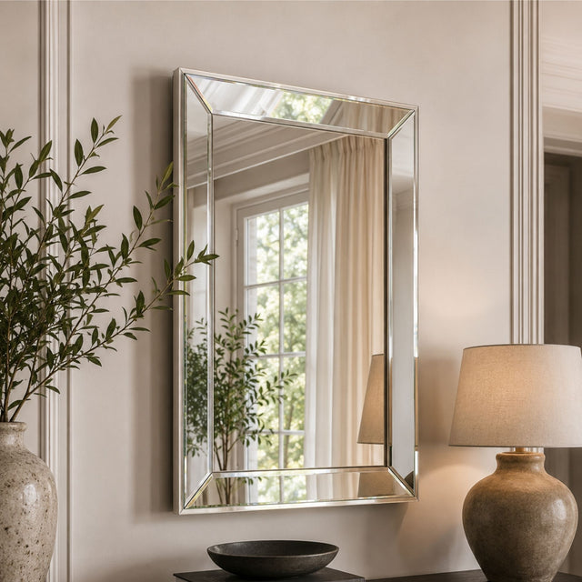 A rectangular wall mirror with a slim silver frame hangs on a beige paneled wall. The mirror reflects a window with cream curtains and greenery, while the surrounding room includes a potted plant on the left and a table lamp on the right.