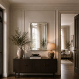 A styled interior entryway featuring a rectangular silver-framed mirror on a dark wood console table, flanked by a textured ceramic lamp and a large vase with greenery. The room has warm beige walls with molding, hardwood floors, and an open doorway revealing a softly lit living area in the background.