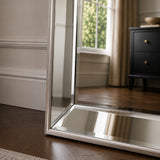 A tall floor mirror with a slim silver metal frame standing on a dark wood floor in a styled room, reflecting a window and a black dresser.