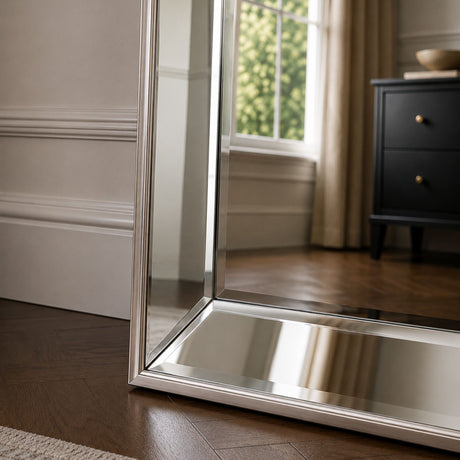 A tall floor mirror with a slim silver metal frame standing on a dark wood floor in a styled room, reflecting a window and a black dresser.