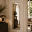 A tall, narrow rectangular wall mirror with a gold-toned frame hangs on a cream paneled wall in a styled interior. The mirror reflects a matching side table and lamp, and the room includes a window, sofa, and decorative plants.
