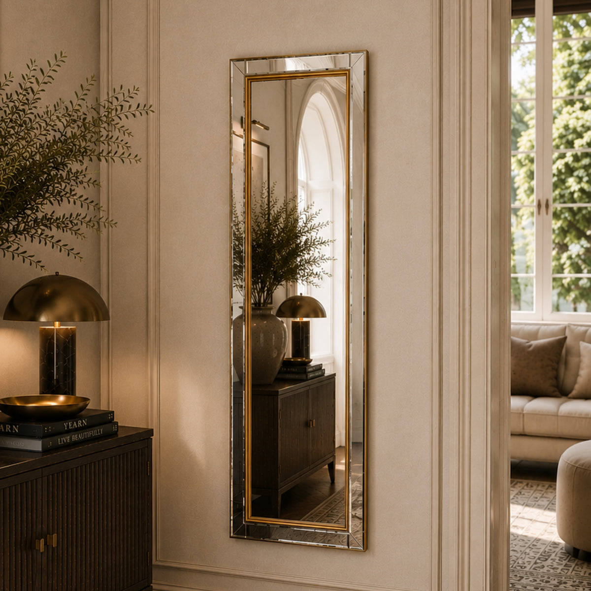 A tall, narrow rectangular wall mirror with a gold-toned frame hangs on a cream paneled wall in a styled interior. The mirror reflects a matching side table and lamp, and the room includes a window, sofa, and decorative plants.