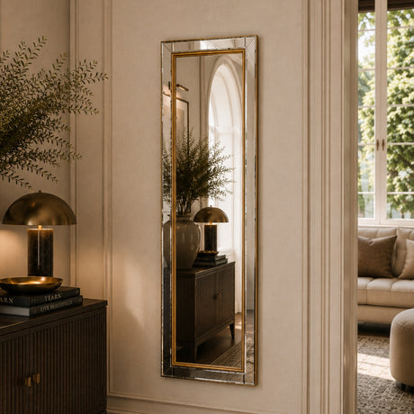 A tall, narrow rectangular wall mirror with a gold-toned frame hangs on a cream paneled wall in a styled interior. The mirror reflects a matching side table and lamp, and the room includes a window, sofa, and decorative plants.