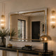 A large rectangular wall mirror with a gold frame hangs above a black console table in a styled interior. The mirror reflects a dark door and room details, while the scene is lit by warm wall sconces and a table lamp.