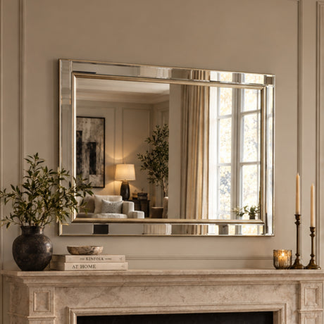 A large rectangular wall mirror with a polished silver frame hangs above a stone fireplace mantel in a styled interior. The mirror reflects a softly lit room with curtains and a window, while the mantel displays a dark vase with greenery, stacked books, and two lit taper candles.