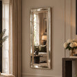 A tall, narrow rectangular wall mirror with a brushed gold-toned frame, mounted on a beige wall in a styled room with curtains, a sofa, and a console table with flowers.