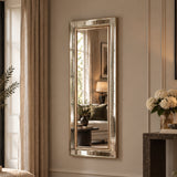 A tall, narrow rectangular wall mirror with a brushed gold-toned frame, mounted on a beige wall in a styled room with curtains, a sofa, and a console table with flowers.