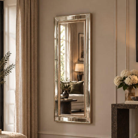 A tall, narrow rectangular wall mirror with a brushed gold-toned frame, mounted on a beige wall in a styled room with curtains, a sofa, and a console table with flowers.