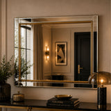 A rectangular wall mirror with a gold-toned frame hangs above a dark console table in a styled interior. The mirror reflects a warmly lit room with curtains, wall sconces, and a dark door.