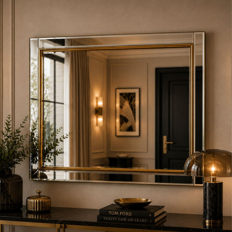 A rectangular wall mirror with a gold-toned frame hangs above a dark console table in a styled interior. The mirror reflects a warmly lit room with curtains, wall sconces, and a dark door.