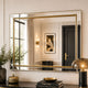 A large rectangular wall mirror with a thin gold frame hangs on a cream wall above a console table. The mirror reflects a classic interior with curtains, a door, and wall sconces.