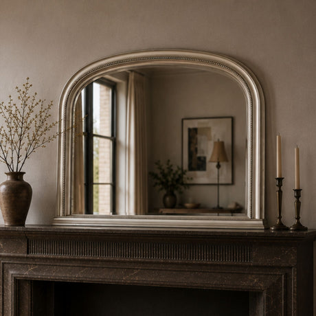 An arched wall mirror with a distressed silver frame resting on a dark fireplace mantel in a styled room. The mirror reflects a softly lit interior window and lamp, while the mantel is decorated with a brown ceramic vase containing dried branches on the left and two dark brass candlesticks with cream candles on the right.