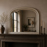 An arched wall mirror with a distressed silver frame resting on a dark fireplace mantel in a styled room. The mirror reflects a softly lit interior window and lamp, while the mantel is decorated with a brown ceramic vase containing dried branches on the left and two dark brass candlesticks with cream candles on the right.