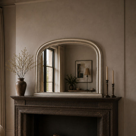 An arched wall mirror with a silver frame mounted above a dark fireplace mantel in a styled room. The mirror reflects a softly lit interior, while the mantel is decorated with a brown vase of dried branches on the left and two black candlesticks on the right.