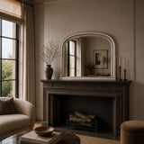 A large arched mirror with a cream frame hangs above a dark fireplace mantel in a styled living room. The mantel holds a tall vase with dried branches and candlesticks, and the room has warm neutral walls, curtains, and a sofa.