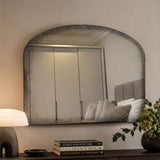 An arched wall mirror with a distressed, textured dark gray frame hangs on a beige wall above a console table. The reflection shows a bedroom interior with a gray headboard and pillows.
