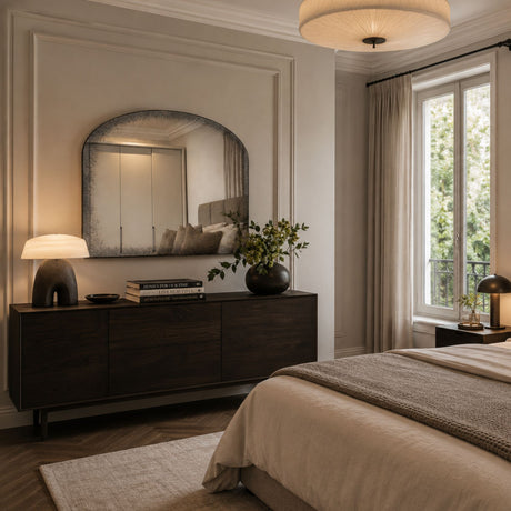 A styled bedroom interior featuring a dark wood dresser with an arched mirror, a lit table lamp, a vase with greenery, and a neatly made bed with beige bedding in a softly lit room with a window and curtains.