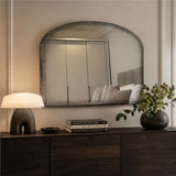 An arched rectangular wall mirror with a distressed, textured dark gray frame hangs above a dark wood console table in a styled bedroom setting. The mirror reflects a bed with gray pillows, and the room is illuminated by a lit table lamp on the left and natural light from the right.