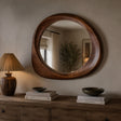 A large oval wooden wall mirror with a thick, rounded handcrafted frame, mounted on a beige wall above a rustic wooden console table with a lamp and books.