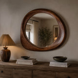 A large oval wooden wall mirror with a thick, rounded handcrafted frame, mounted on a beige wall above a rustic wooden console table with a lamp and books.