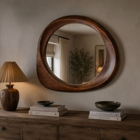 A large oval wooden wall mirror with a thick, rounded handcrafted frame, mounted on a beige wall above a rustic wooden console table with a lamp and books.