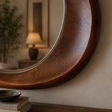 A close-up of a large round wall mirror with a thick, curved dark brown wooden frame, mounted on a beige wall above a wooden console table.