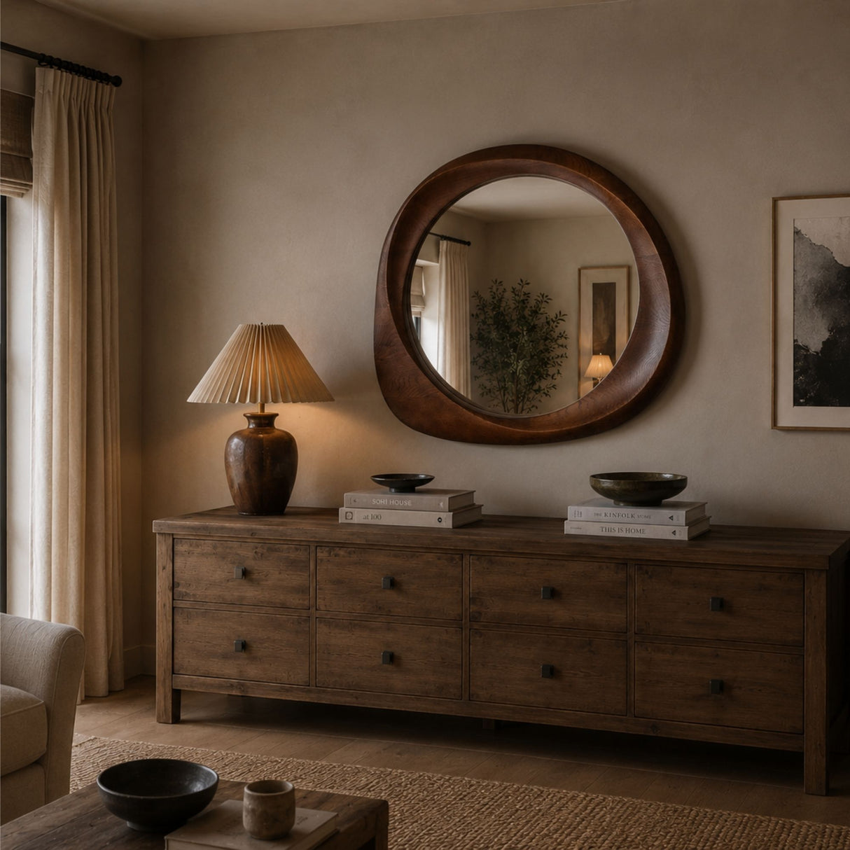 A large round wooden wall mirror hangs above a rustic wood dresser in a warmly lit living room. A lit table lamp, stacked books, and dark bowls sit on the dresser, with a chair and table partially visible in the foreground.