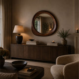 A round wooden-framed wall mirror hangs above a dark wood sideboard in a warmly lit living room. A table lamp, books, a vase with greenery, and a chair are visible in the styled interior.