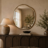 An organic-shaped round wall mirror with a gold-toned frame hangs above a dark wood console table in a warm, neutral-toned room. The mirror reflects a softly lit interior window, while the table is styled with a textured ceramic table lamp, a black bowl, a book, and a vase with dried branches.