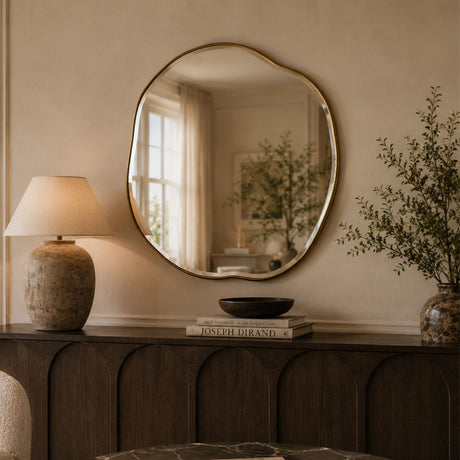 An organic-shaped round wall mirror with a gold-toned frame hangs above a dark wood console table in a warm, neutral-toned room. The mirror reflects a softly lit interior window, while the table is styled with a textured ceramic table lamp, a black bowl, a book, and a vase with dried branches.