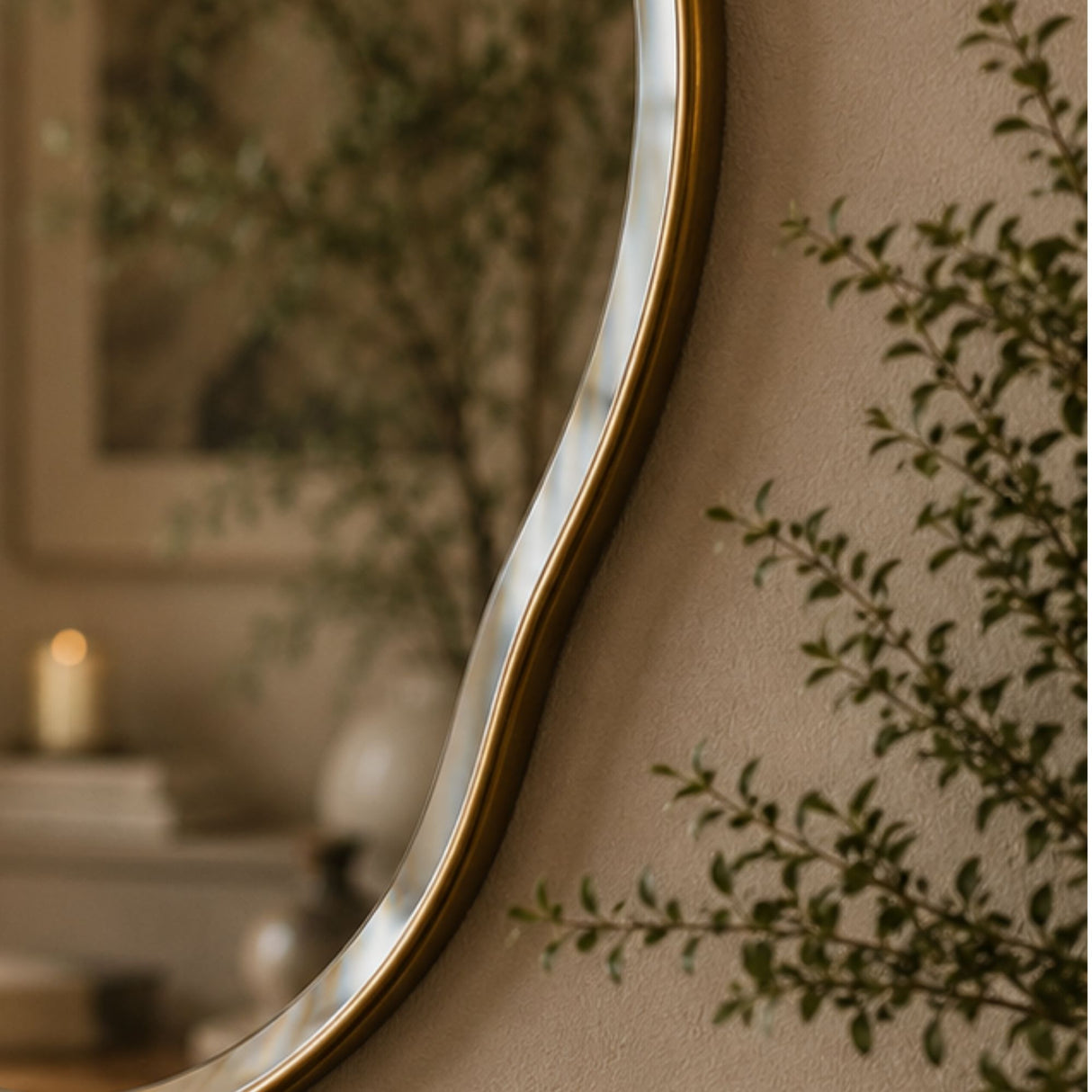 A close-up of a decorative wall mirror with a wavy organic border and a warm metallic gold frame, mounted on a beige wall with soft-focus greenery and a candle in the background.