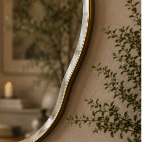 A close-up of a decorative wall mirror with a wavy organic border and a warm metallic gold frame, mounted on a beige wall with soft-focus greenery and a candle in the background.