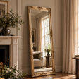 A tall floor-length mirror with an ornate gold frame stands leaning against a cream paneled wall in a sunlit living room. The mirror reflects a window, curtains, and a light sofa, while the room itself includes a fireplace, plants, and draped curtains.