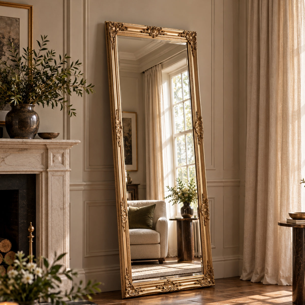 A tall floor-length mirror with an ornate gold frame stands leaning against a cream paneled wall in a sunlit living room. The mirror reflects a window, curtains, and a light sofa, while the room itself includes a fireplace, plants, and draped curtains.