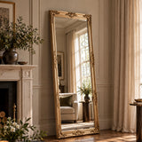 A tall floor-length mirror with an ornate gold frame stands leaning against a cream paneled wall in a sunlit living room. The mirror reflects a window, curtains, and a light sofa, while the room itself includes a fireplace, plants, and draped curtains.
