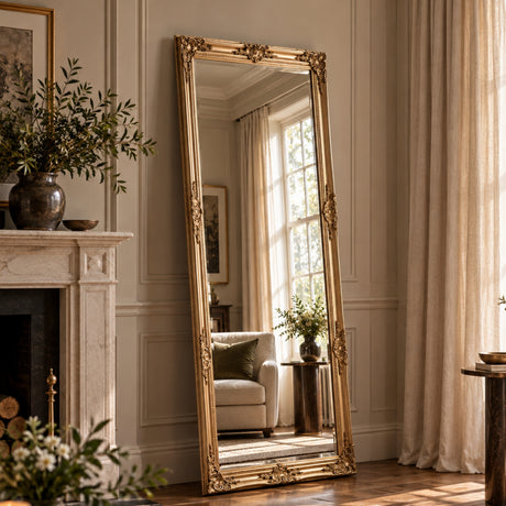 A tall floor-length mirror with an ornate gold frame stands leaning against a cream paneled wall in a sunlit living room. The mirror reflects a window, curtains, and a light sofa, while the room itself includes a fireplace, plants, and draped curtains.