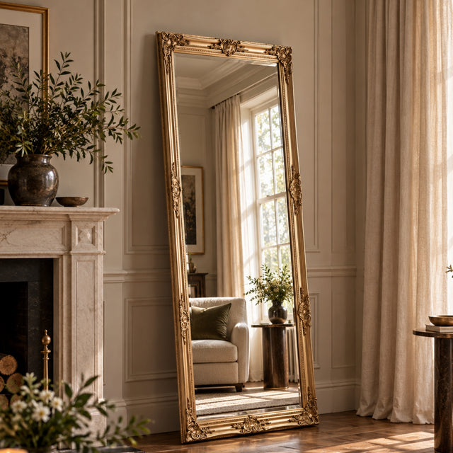 A tall floor-length mirror with an ornate gold frame stands leaning against a cream paneled wall in a sunlit living room. The mirror reflects a window, curtains, and a light sofa, while the room itself includes a fireplace, plants, and draped curtains.