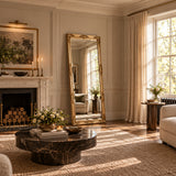 A luxury full-length floor mirror with an ornate gold frame stands in a beautifully styled living room next to a white marble fireplace and a large window with beige curtains.