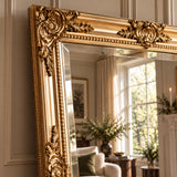 An ornate gold-framed mirror with intricate carved floral and scroll details, leaning against a paneled wall in a styled room with a window, curtains, and home decor visible in the reflection.