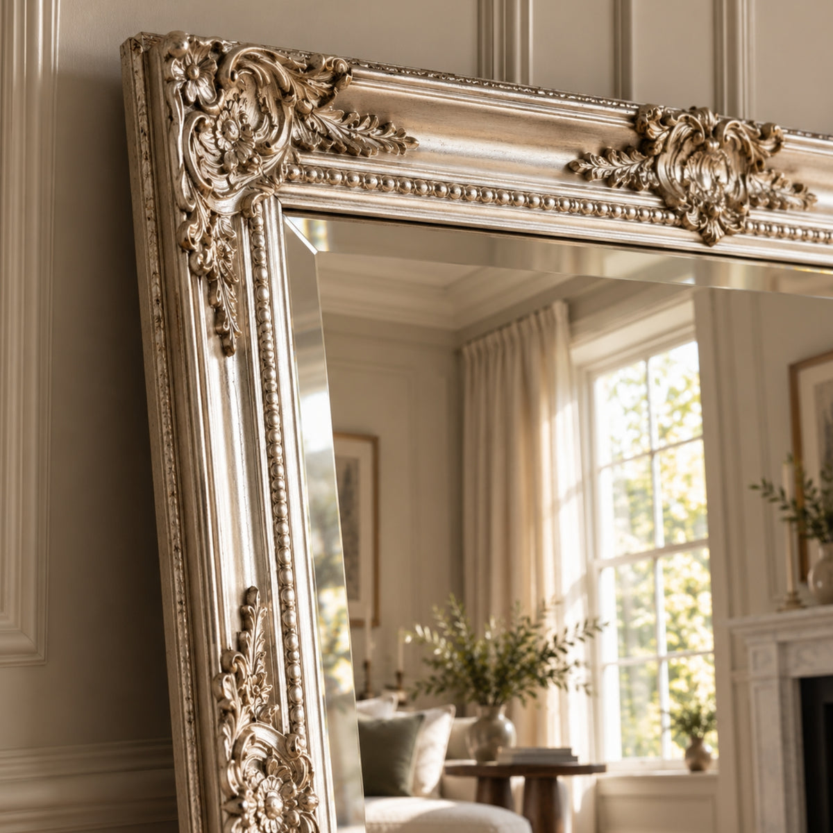 An ornate gold-framed floor mirror leaning against a beige paneled wall, reflecting a softly lit living room interior with curtains, a window, and plants.
